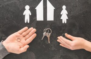 illustrative photo of paper house split in half with paper dolls beside it and two hands holding rings on either side